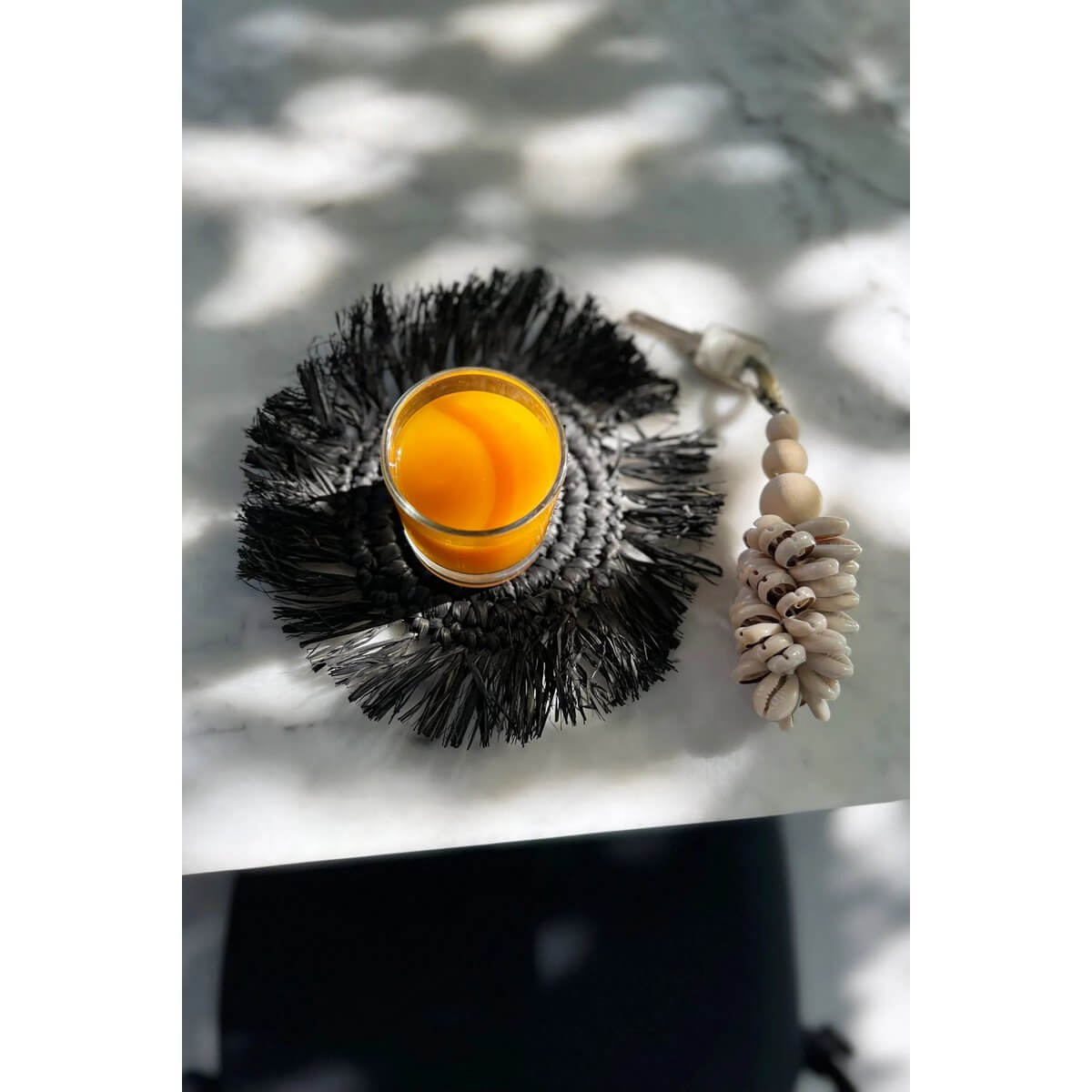 Bohemian black fringe raffia coaster by Bazar Bizar with orange drink on white table, ideal for stylish interiors and exteriors.