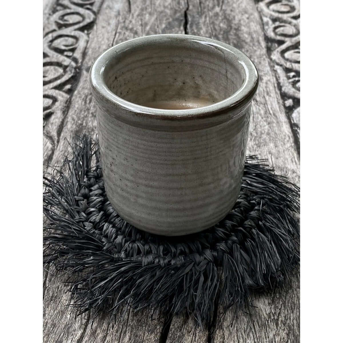 Bohemian Fringe Raffia Coaster in zwart color under a ceramic cup, perfect for stylish interiors by Bazar Bizar Living.