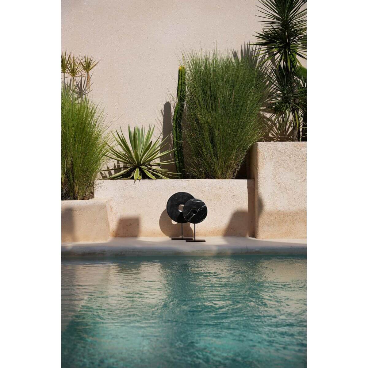 Zwarte marmeren schijf op standaard bij het zwembad, omringd door groene planten, voor een elegante buitenruimte.