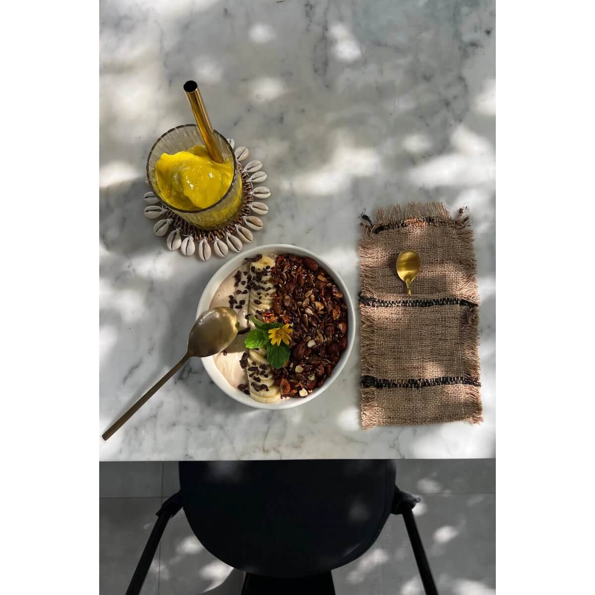 Bohemian style table setting with a beige and black cutlery holder, smoothie, and granola bowl on a marble table surface.