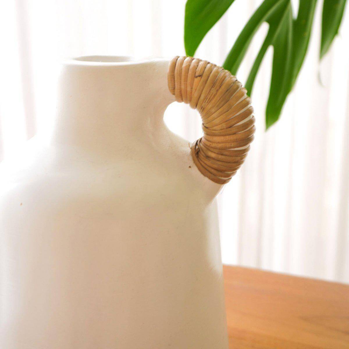Handgemaakte witte vaas met rieten handvat op houten tafel, omgeven door groene planten. Ideaal voor gedroogde bloemen in bohostijl.