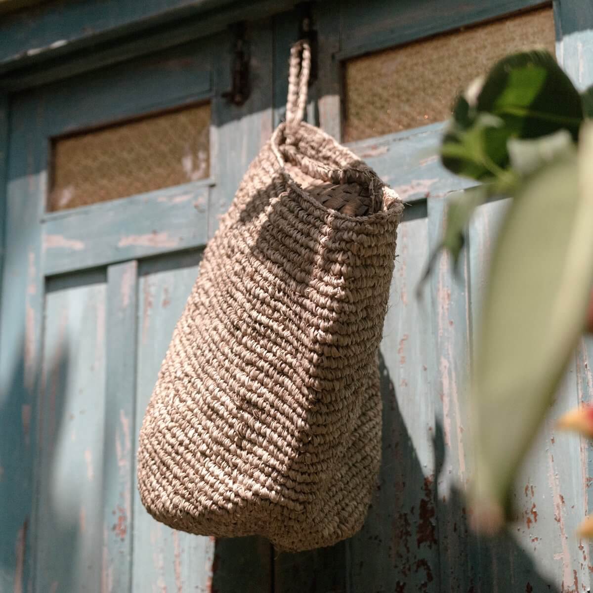 Raffia wandmand UTARA, handgemaakt, hangend aan blauwe rustieke deur, duurzame en praktische opbergoplossing.