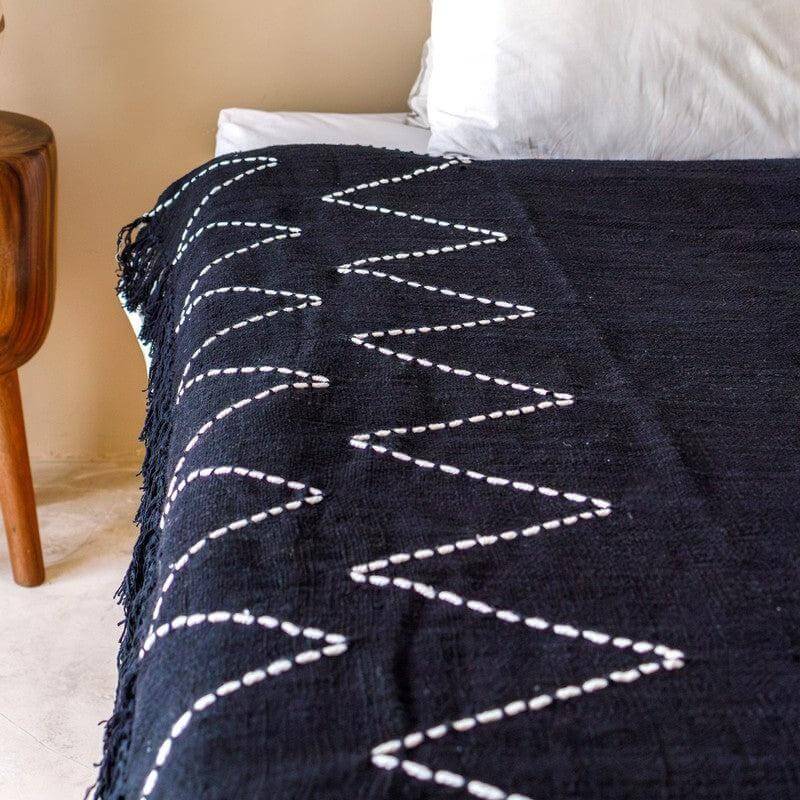 Boho Deken Zwart Handgeweven Katoenen Deken 140x200 cm met wit zigzagpatroon, op bed naast houten nachtkastje