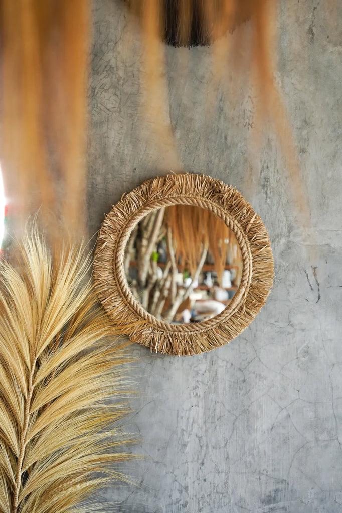 Boho spiegel The Tahiti in Indonesische raffia tegen een grijze muur met decoratieve bladeren erbij.