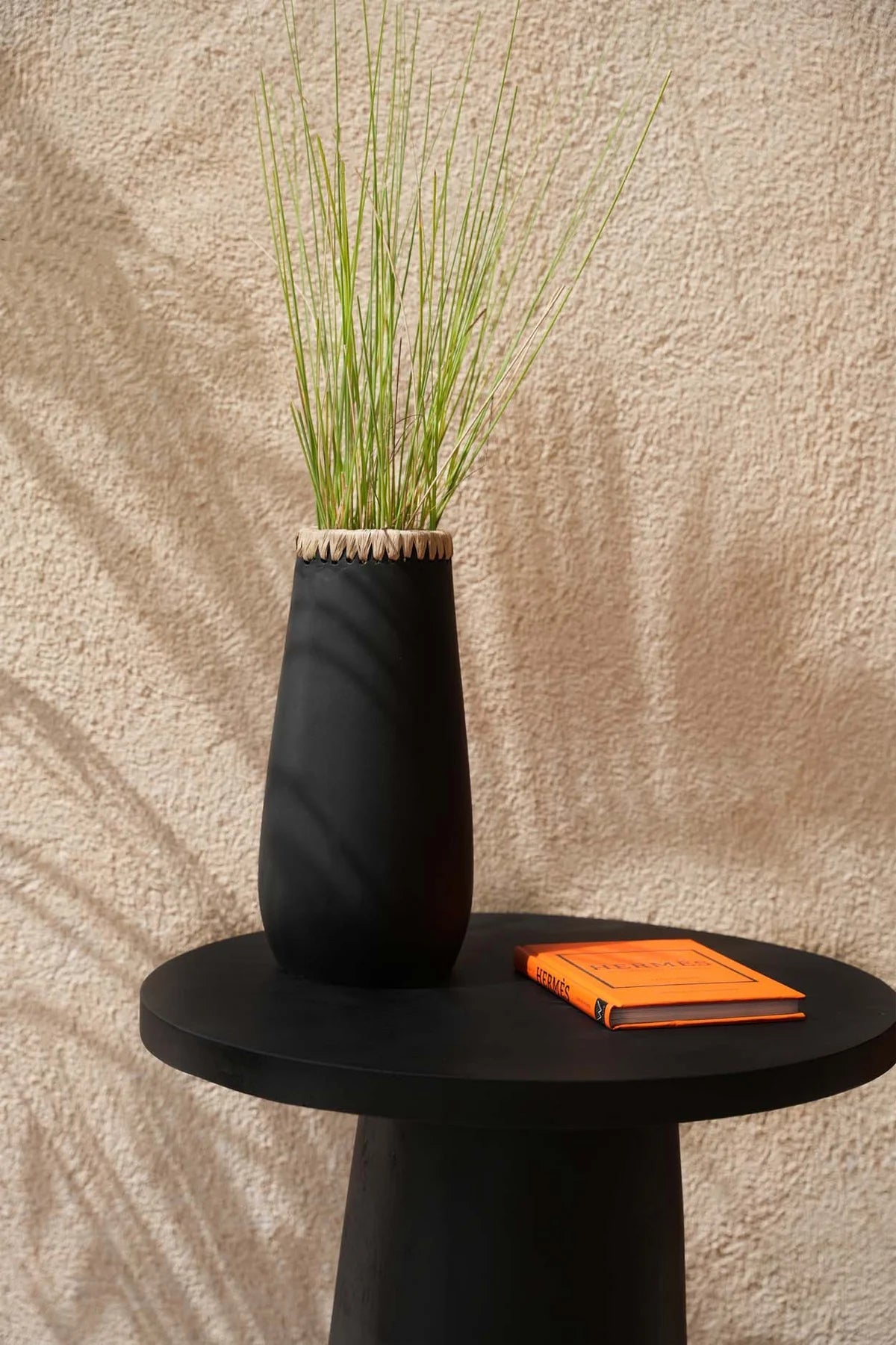 Zwarte Boho vaas The Sneaky L op ronde tafel, elegant handgemaakt design met groene plantenstengels, perfecte aanvulling voor modern interieur.