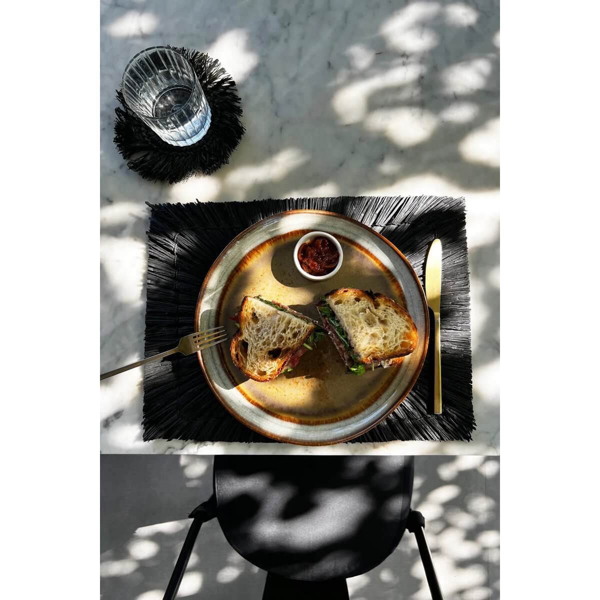 Stylish black bohemian placemat with sandwich and glass on a sunlit table.