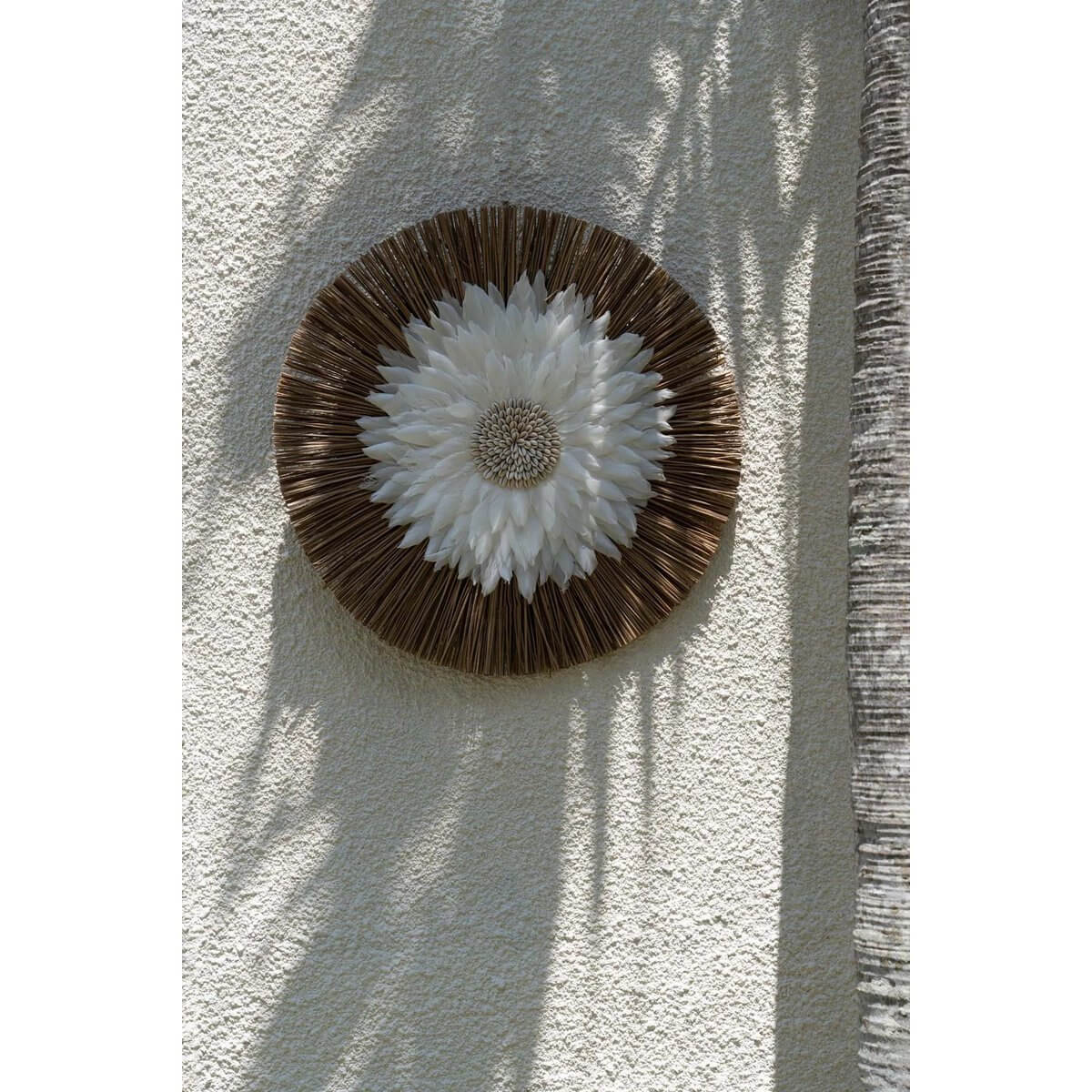 Rond Alang Feather Juju kunstwerk met witte veren en kaurischelpen op droog gras tegen een beige muur.
