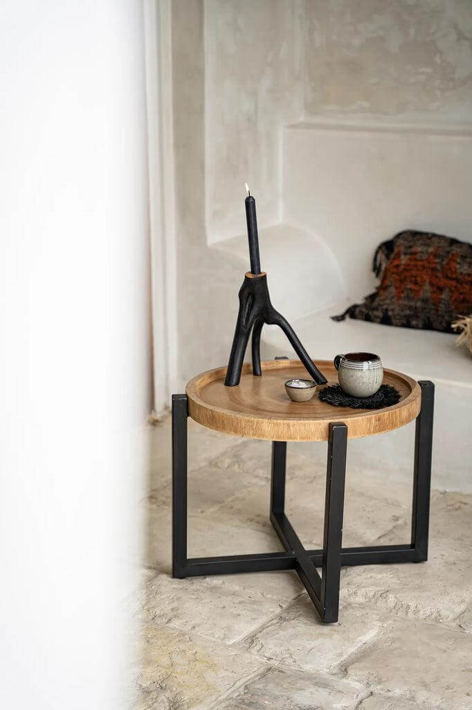 Ronde bijzettafel met houten blad en zwarte poten, gedecoreerd met een zwarte kaars en een koffiebeker op een beige vloer.