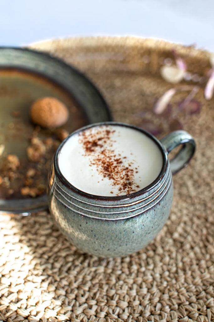 Keramische koffiemok gevuld met schuimende koffie en een snufje kaneel, omringd door een rustieke setting op een gevlochten ondergrond.