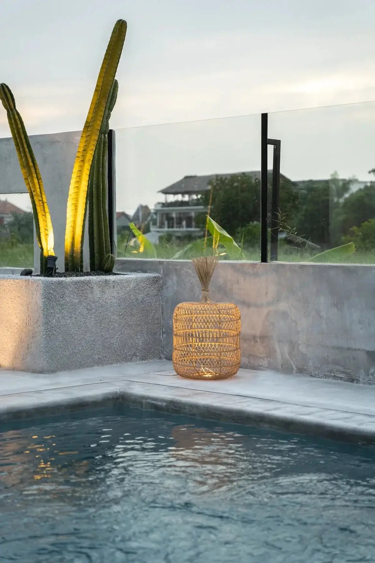 Sfeervolle rotan lamp naast zwembad met cactussen in een tuin bij zonsondergang.