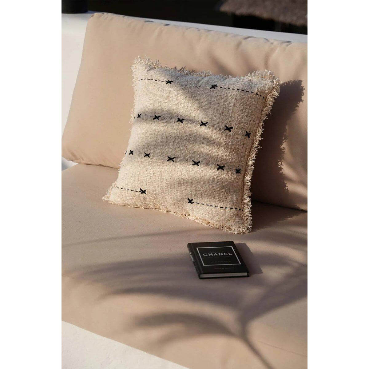 Natuurlijke zwarte French Kiss kussenhoes op bank met boek, creëert Parijse charme in huis, perfect voor woonkamer en slaapkamer.
