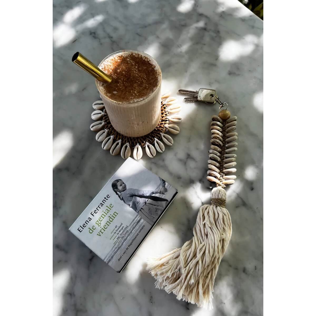Bohemian macrame fishbone keychain with shells and tassel, next to a drink on a shell coaster and a book.