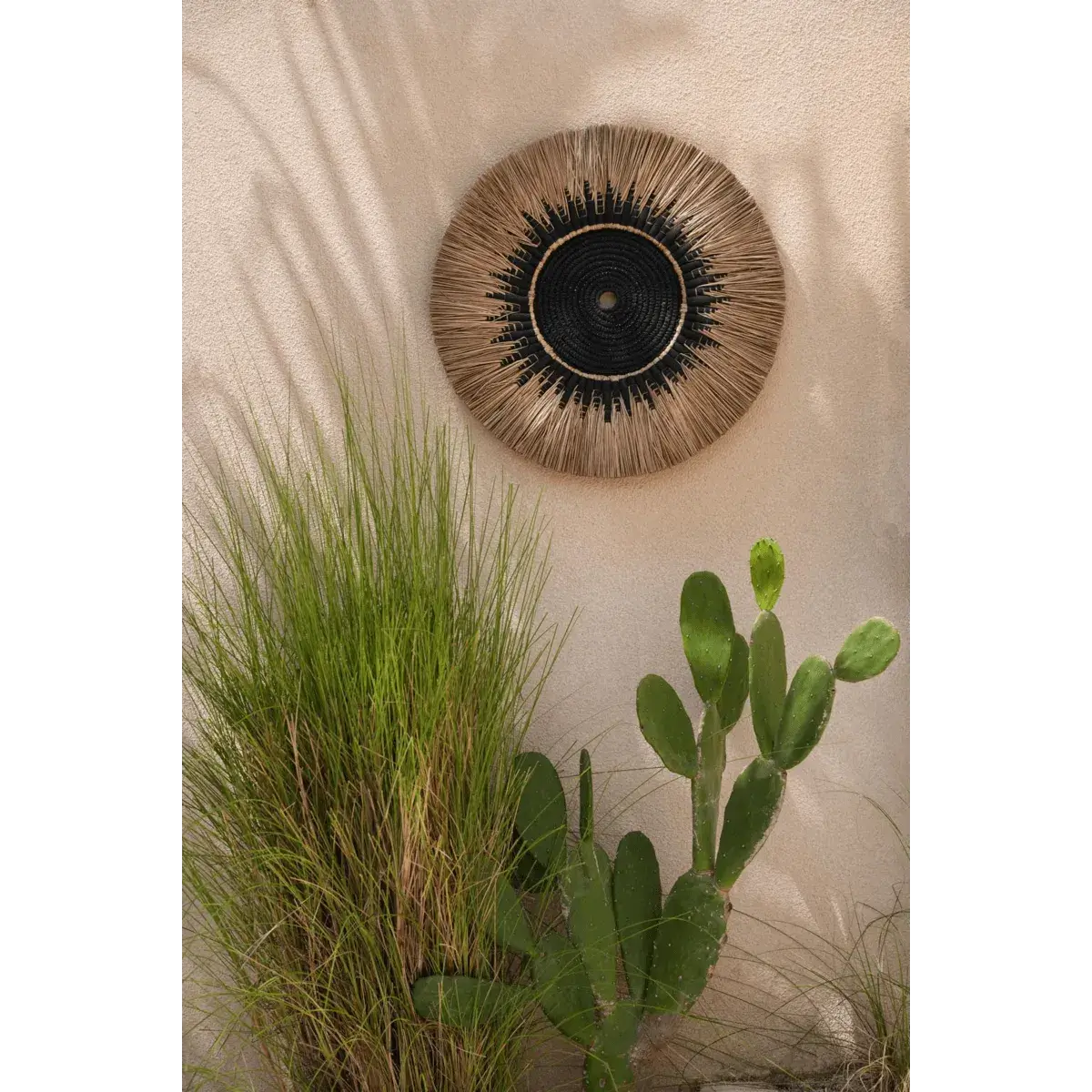 Bohemian black and natural raffia macramé wall decor by Bazar Bizar Living, adding a coastal vibe next to green outdoor plants.