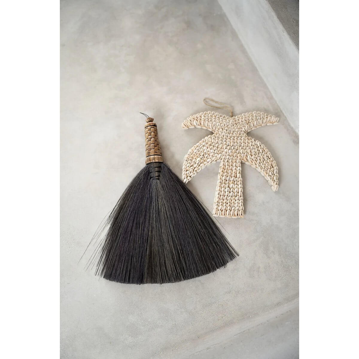 Bohemian palm-shaped shell decor piece handmade in Bali, alongside a traditional broom on a concrete background. Perfect for home styling.
