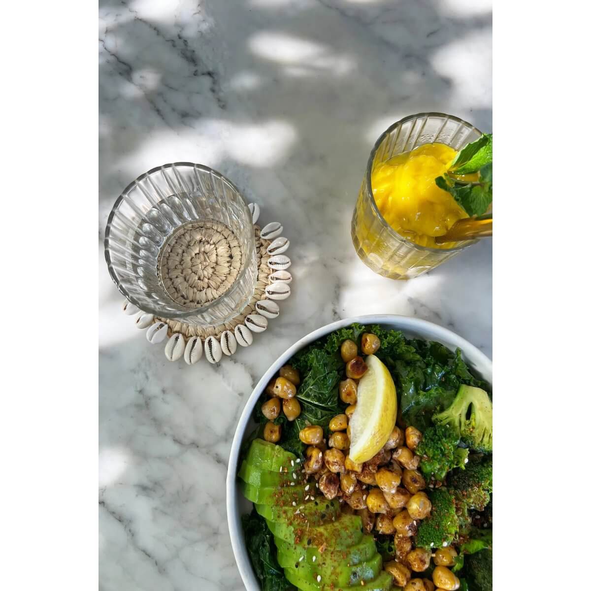 De Seagrass Shell Coaster under glass on marble table with salad and lemon drink, showcasing Bohemian island style.