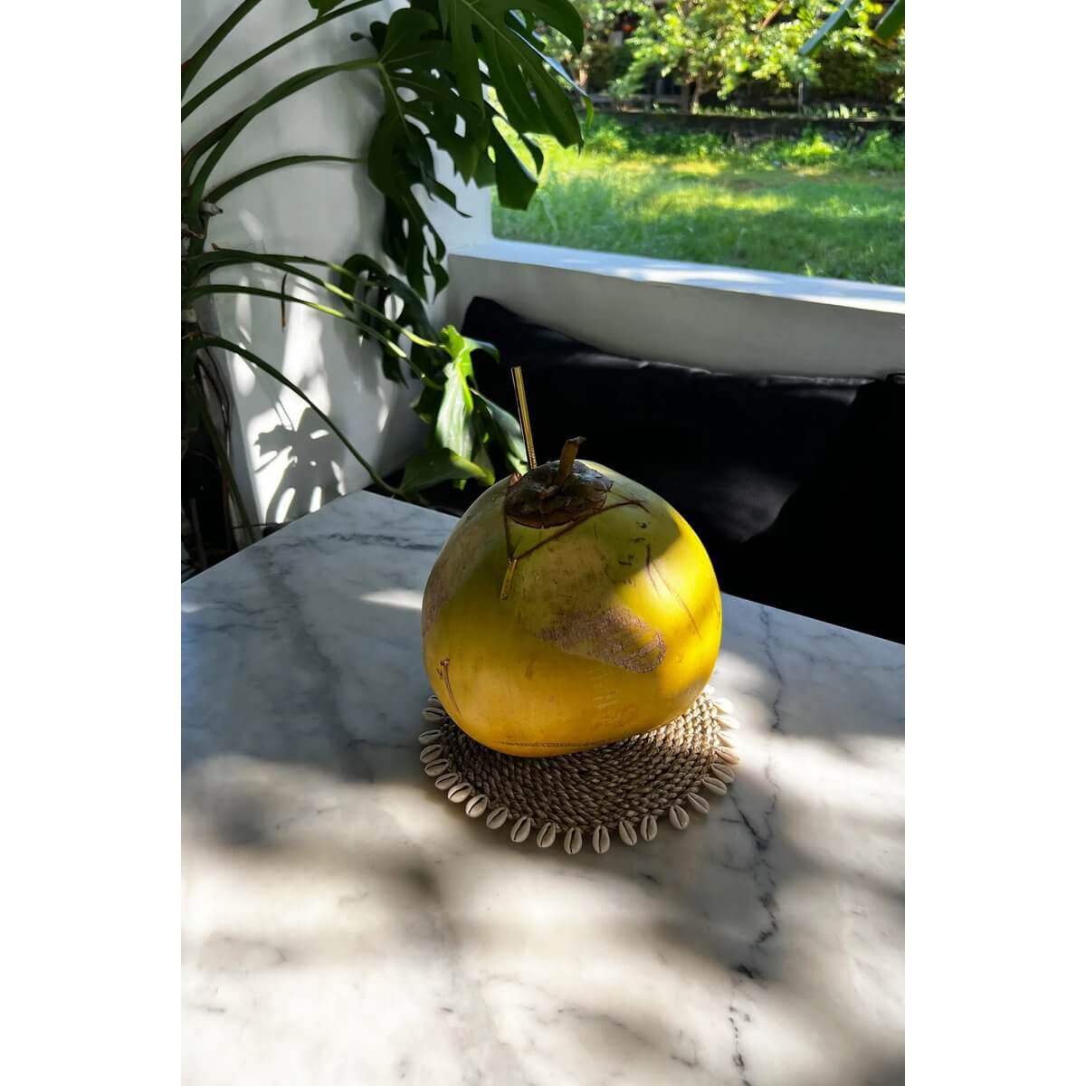 Gele kokosnoot op een zeegras onderzetter met schelpen, op een buitenmarmeren tafel.