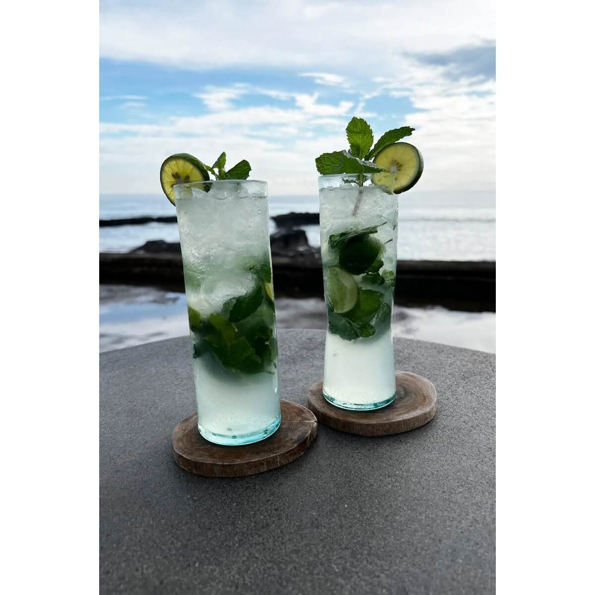 Two mojito cocktails in tall glasses on teak root boho coasters by the ocean, refreshing summer vibes.