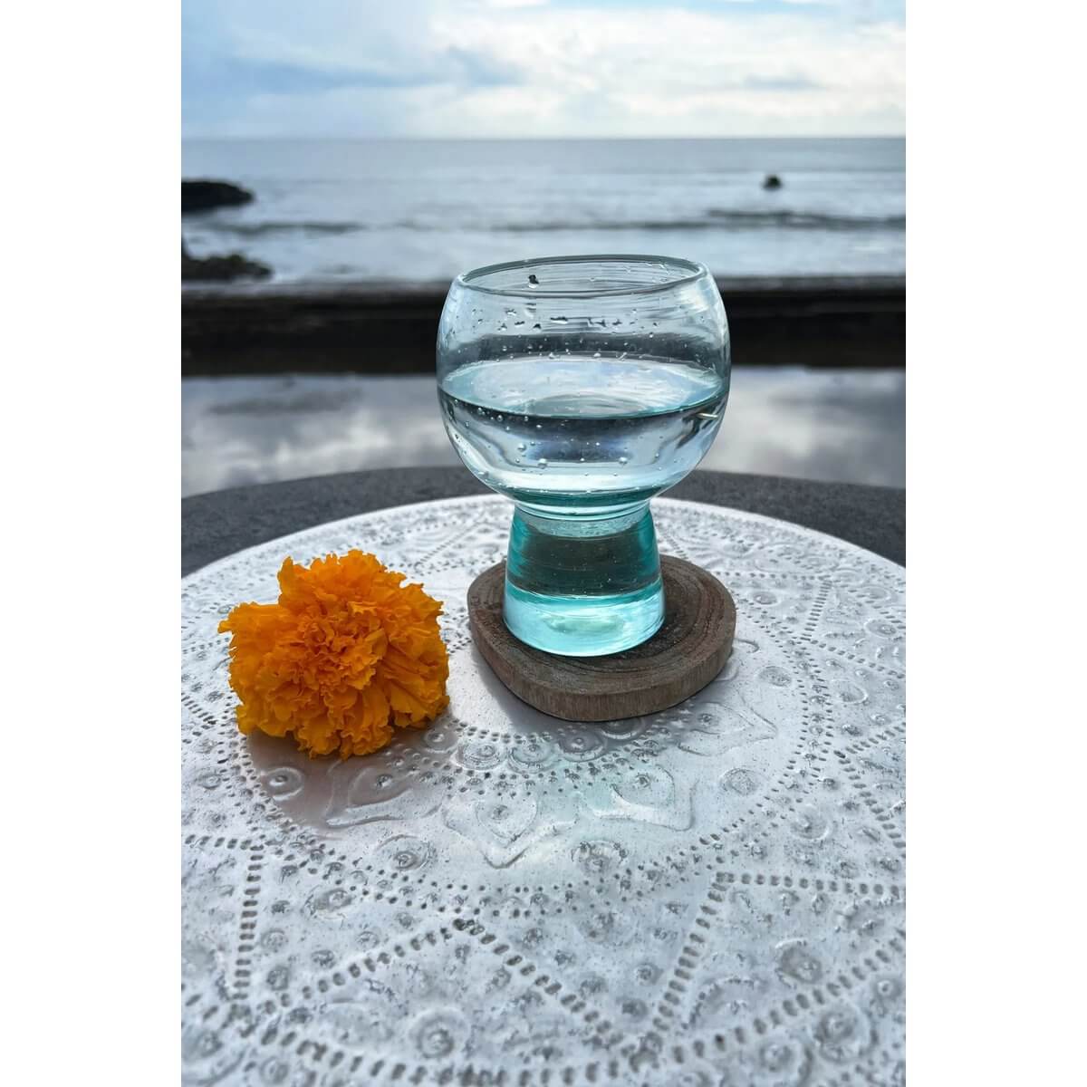 Glass of water on a teak root boho coaster with a marigold flower against a scenic ocean backdrop, perfect for bohemian decor.