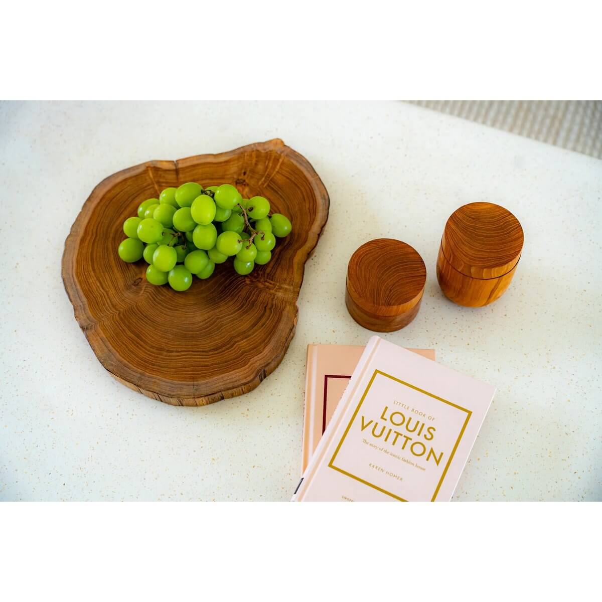Rustic bohemian setup with green grapes on a teakwood slab, small teak root jars, and a fashion book on a white table.