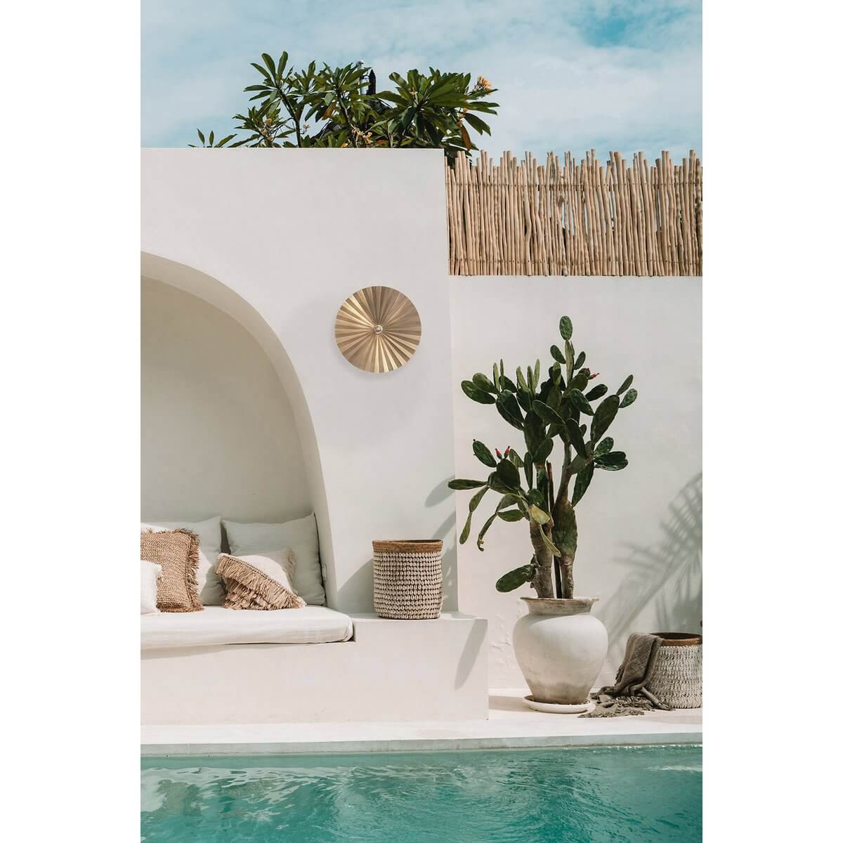 Bohemian outdoor seating area with wall-mounted brass decoration, large potted cactus, and white cushions by a poolside.