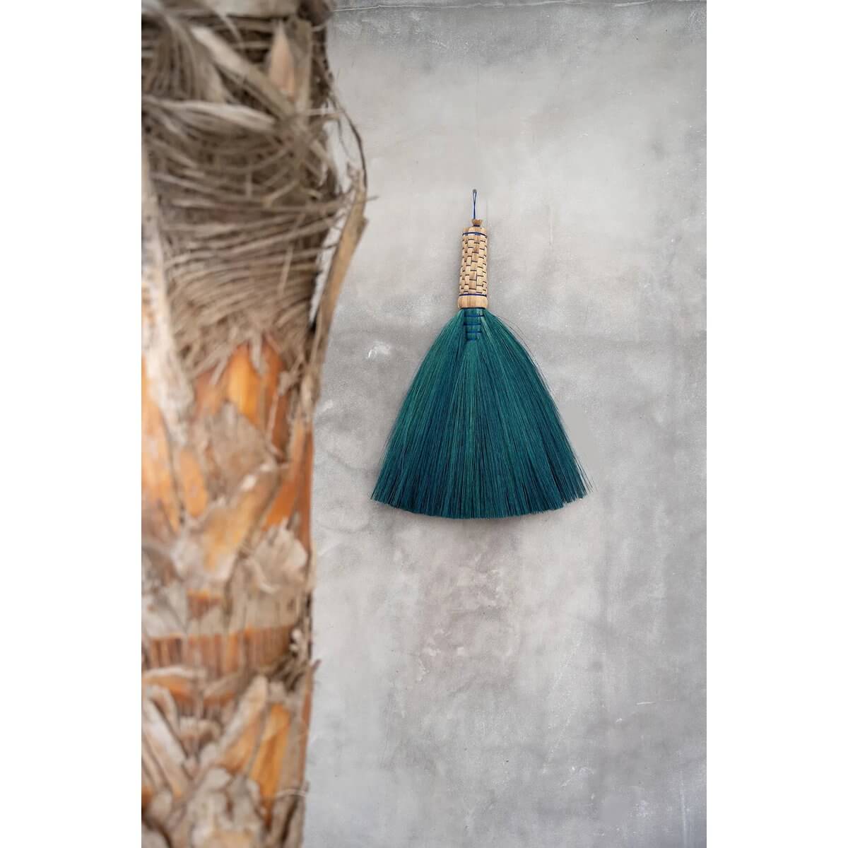 Alt Tag: Turquoise Indonesian grass hand broom, a Bohemian home accessory for clearing dust, elegantly displayed against a textured wall.