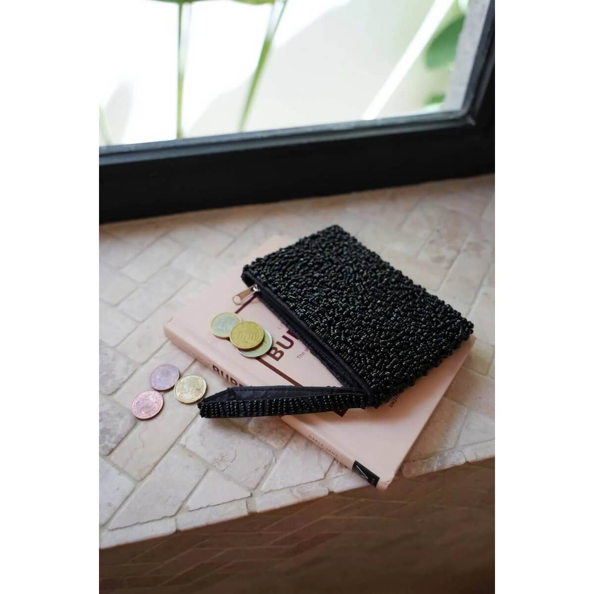 Elegant black beaded wallet with bohemian charm, placed on a book beside coins near a window.
