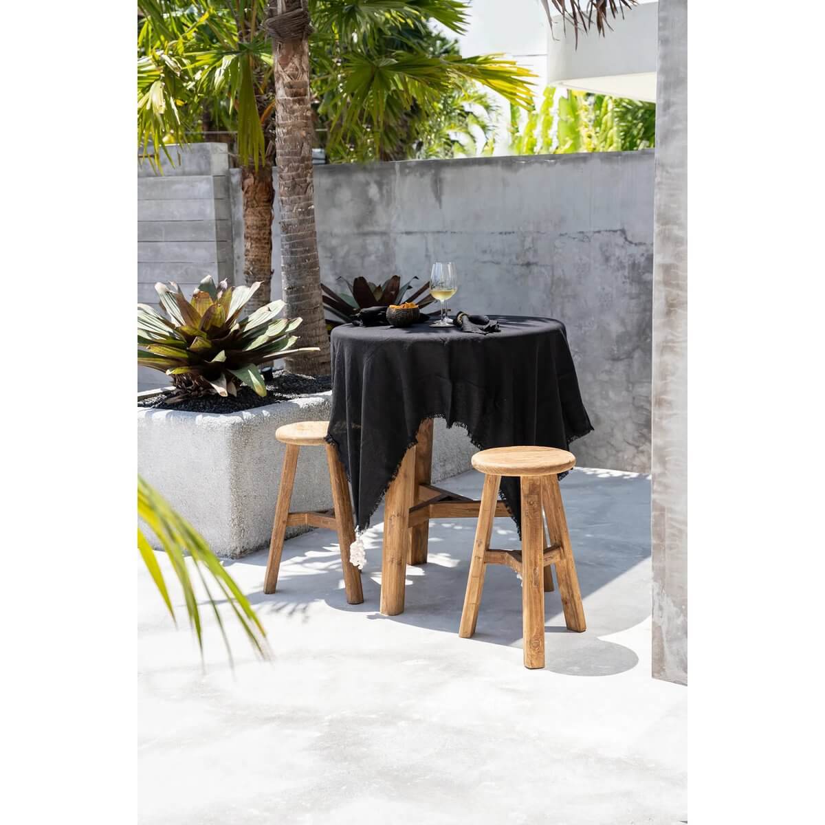 Zwart linnen tafelkleed van 150x150 cm, buiten gedekt op een ronde tafel met houten krukken, omringd door tropische planten.