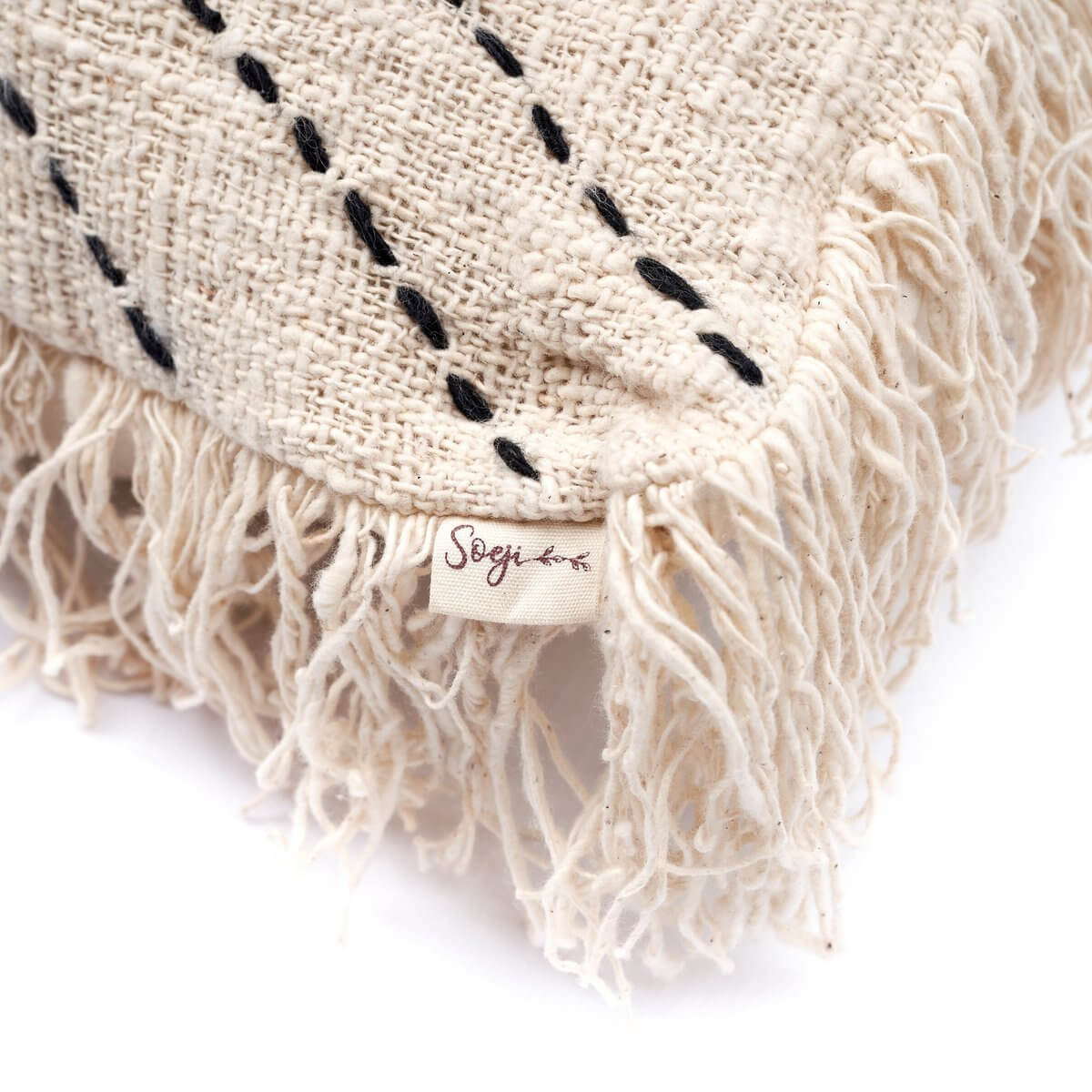 Close-up of a handwoven bohemian decorative pillow cover with fringe detail, made of natural cotton. Tag displaying brand name.