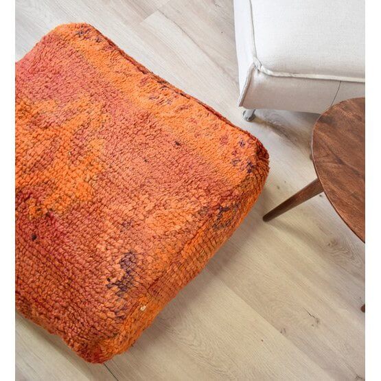 Rood-oranje Marokkaans vloerkussen van vintage Berberkleed in bohemian interieur, naast houten tafel en witte stoel.