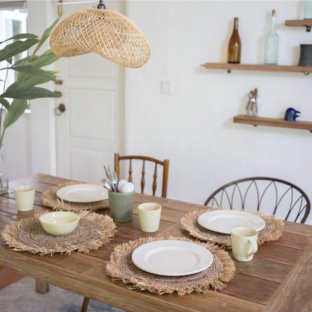 Rotanlamp Padma hangend boven houten eettafel met rustieke placemats, creëert warme en rustgevende lichtpatronen in de eetkamer.
