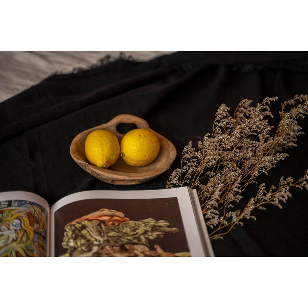 Teakwood bowl with lemons, dried florals, and an open book, exuding a Bohemian vibe on a dark fabric backdrop.