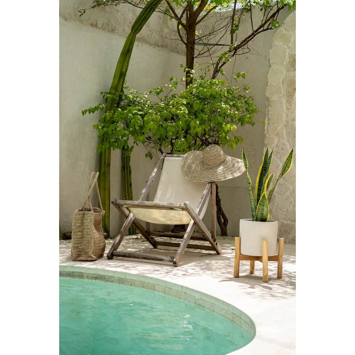 Bohemian garden retreat with lounge chair, woven hat, plant in terracotta pot on teak stand, and serene green pool.