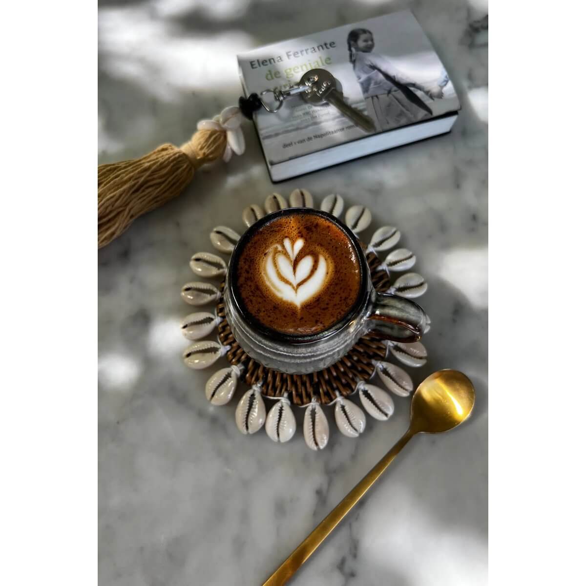 Bohemian coffee scene with a cappuccino on a shell-decorated coaster, gold spoon, and book on a marble table.
