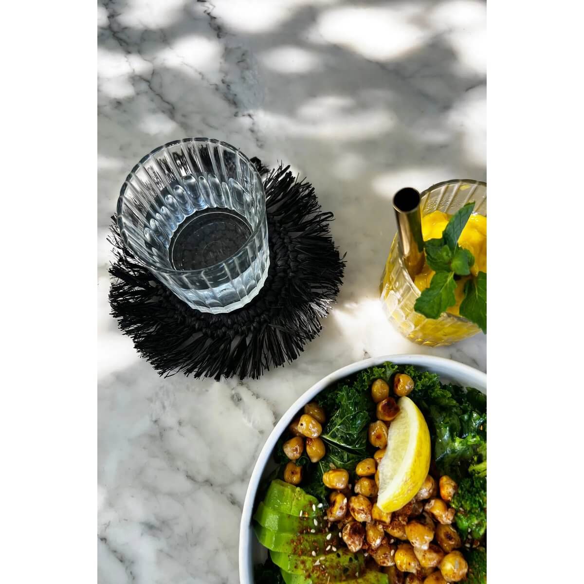 Bohemian Fringe Raffia Coaster in black holding a glass, paired with fresh salad on marble table, by Bazar Bizar Living.