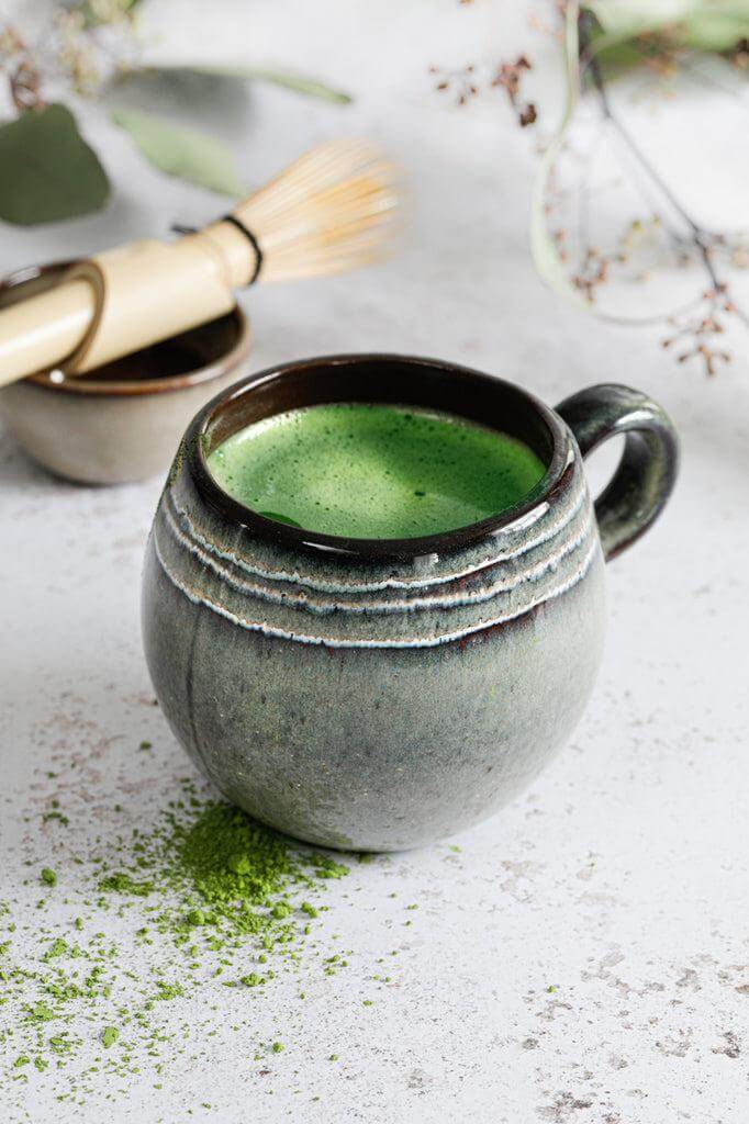 Keramieken koffiemok met matcha thee op rustieke tafel, omringd door een Japanse thee kwast en matchapoeder.