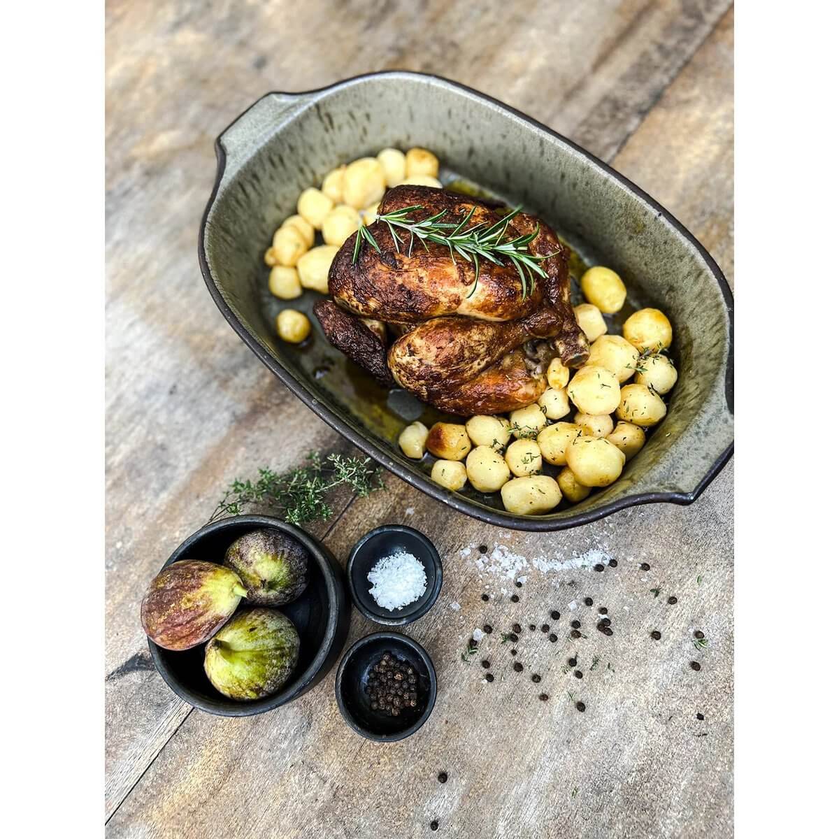 De Comporta Ovenschaal met gebraden kip, aardappelen en kruiden, stijlvol gepresenteerd op houten tafel.