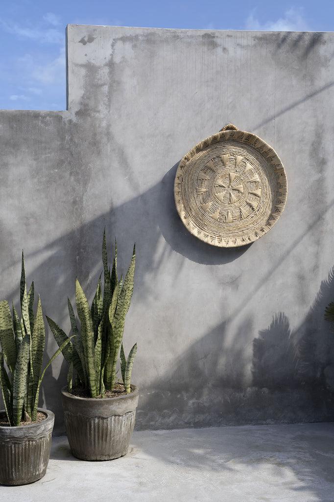 De Xolo muurdecoratie van bananenblad en rotan op een betonnen muur, omringd door groene planten, in natuurlijke tinten.