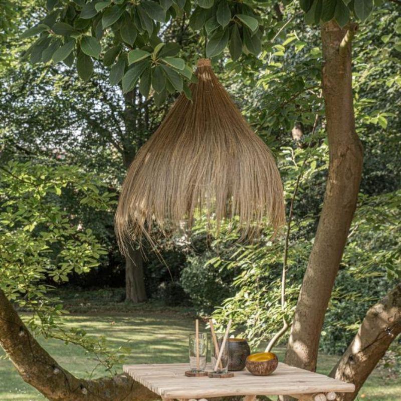 Hanglamp Rayung Hut hangt aan een boom, omgeven door groen, creëert een bohemien sfeer met duurzaam Rayung Gras.