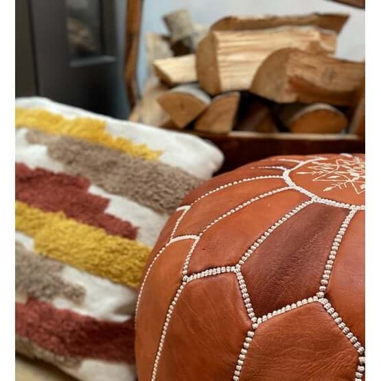 Handgemaakte Marokkaanse terrabruine leren poef naast kleurrijk kussen en houtblokken, perfect voor een boho sfeer in huis.